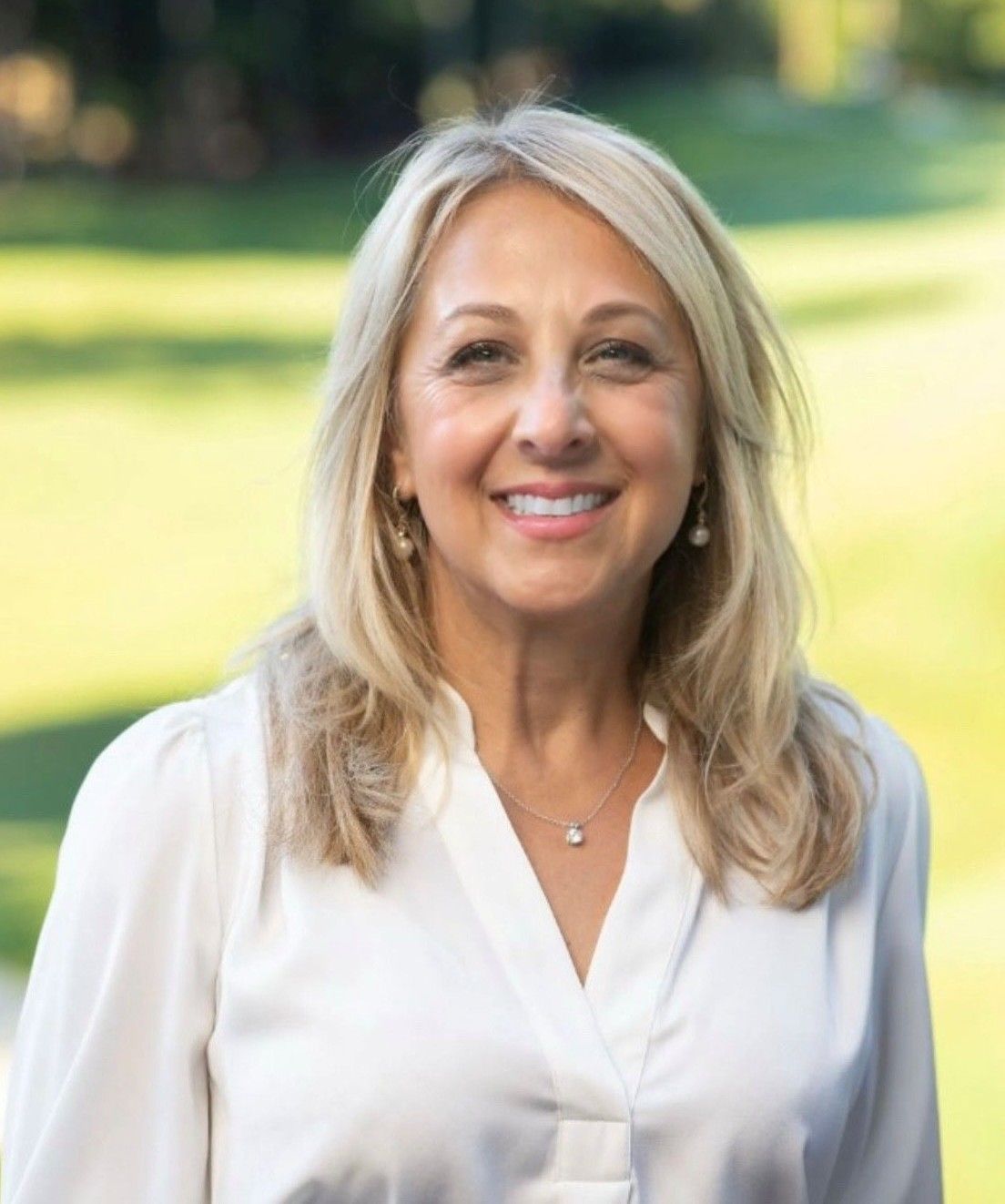 Woman with blonde hair smiling, wearing a white blouse, outdoors. Green grass and trees are in the background.
