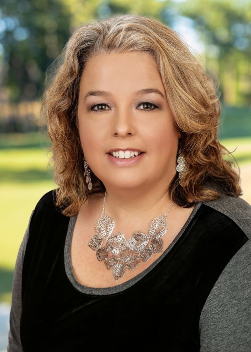 Woman with blonde hair smiling, wearing a black and grey top, and a silver necklace against a brick wall background.