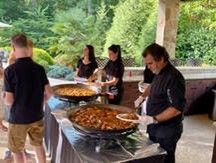 People serving food from large paella pans at an outdoor event. Several servers in black are visible.