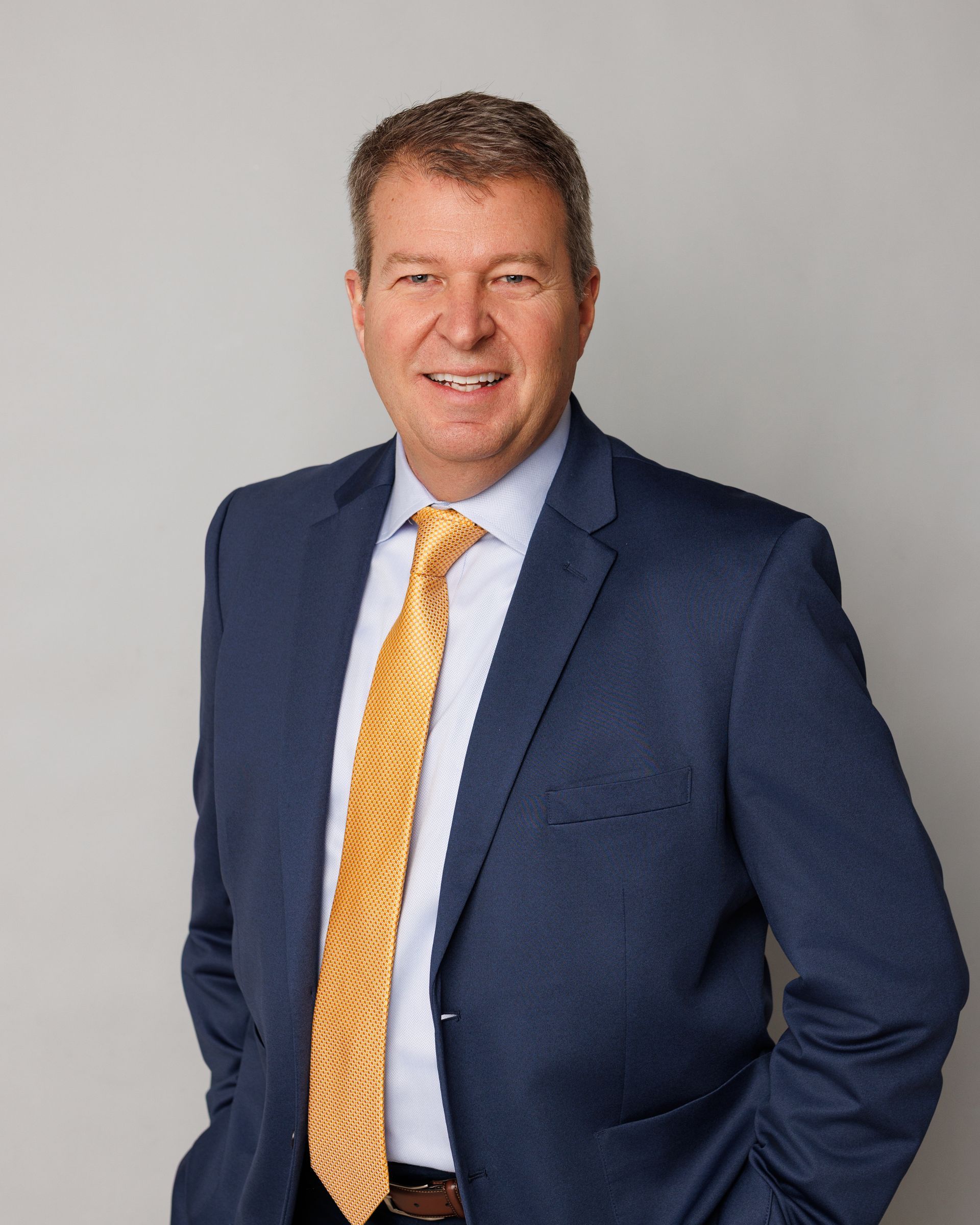 A man in a suit and white shirt is smiling in front of a brick wall.