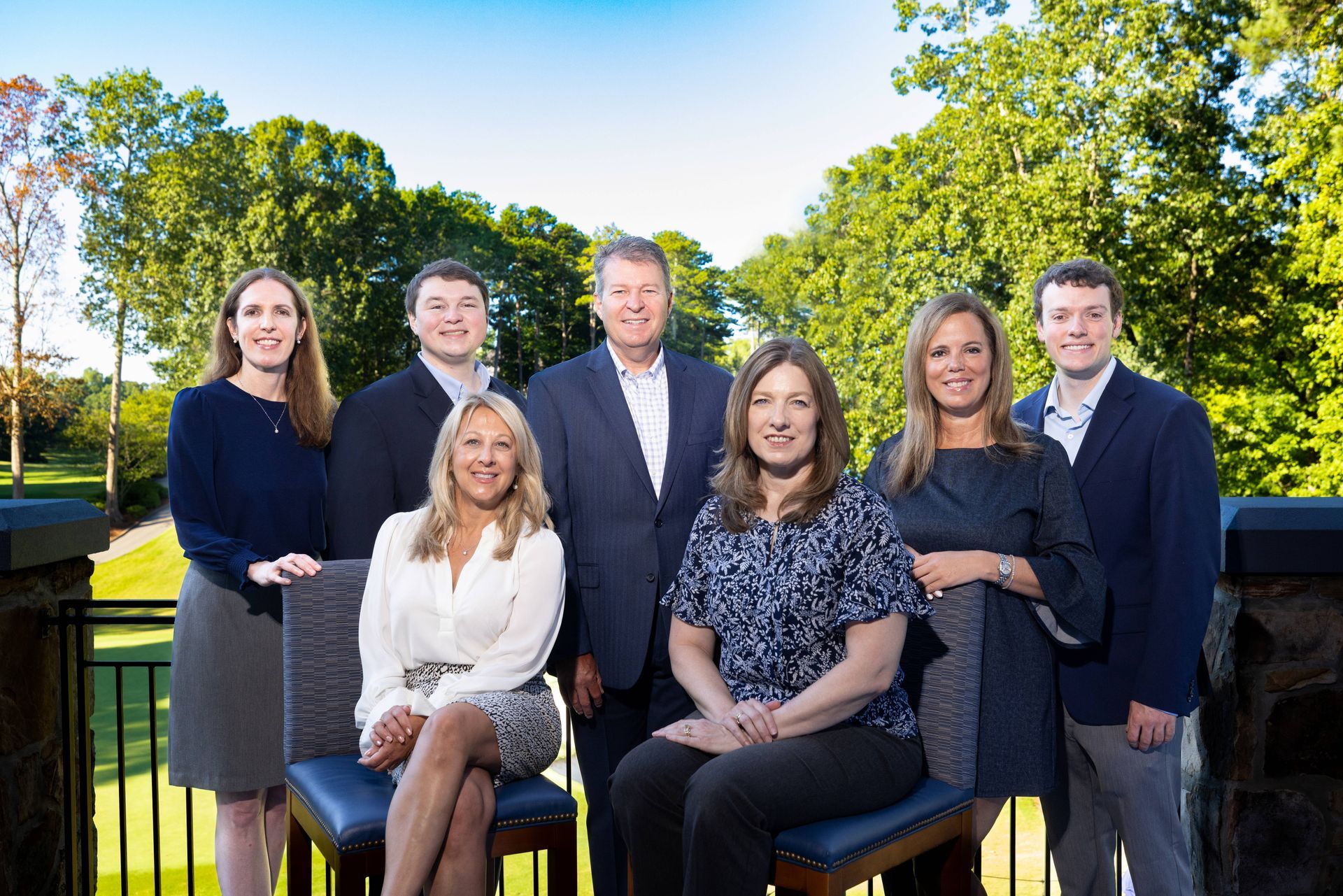 Group of nine professionals posing outdoors; some seated, some standing; trees and golf course in background.