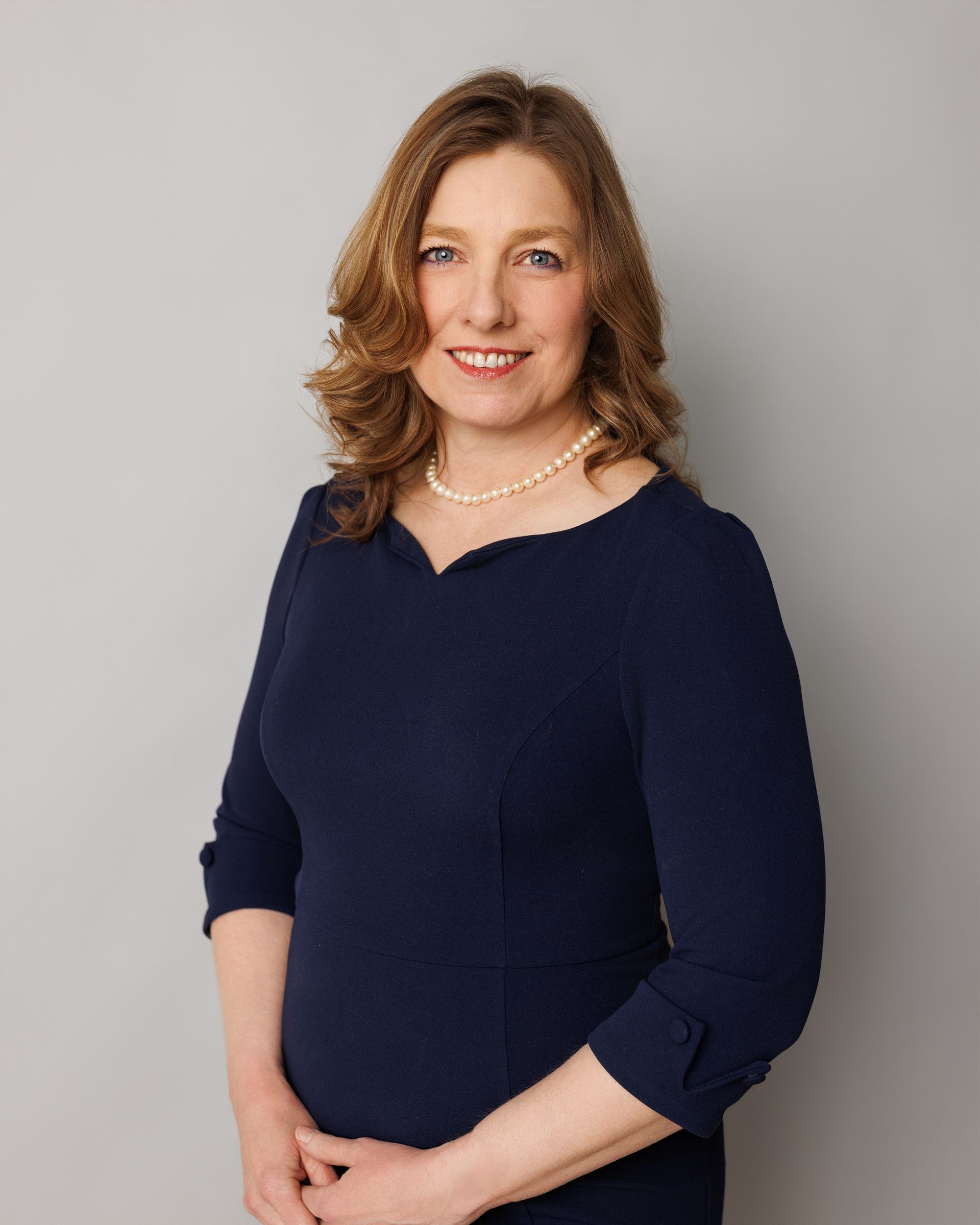 Smiling woman with light brown hair, wearing a navy top, stands in a green, outdoor setting.