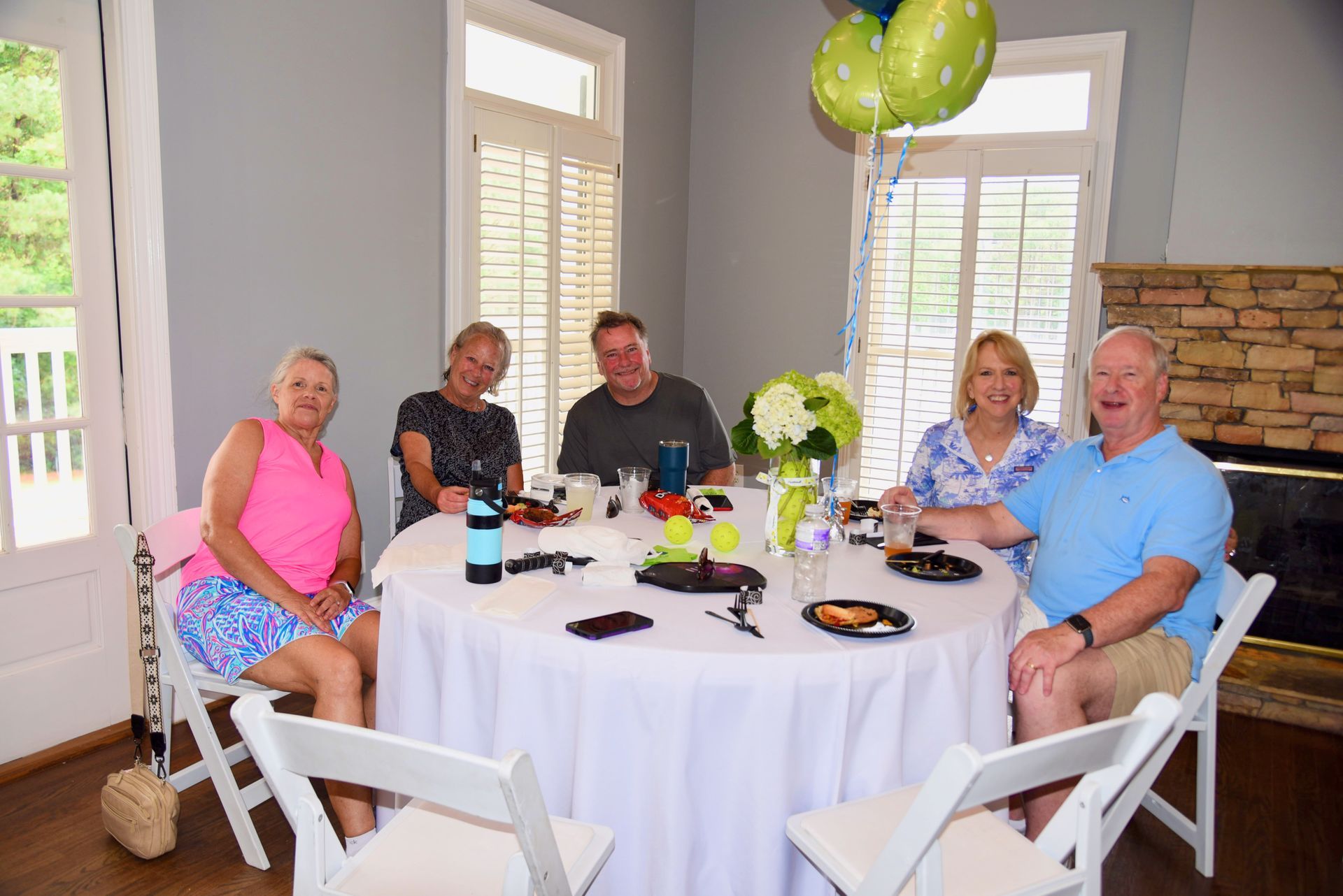 A group of six people seated around a round table set for a meal, with balloons in the background.