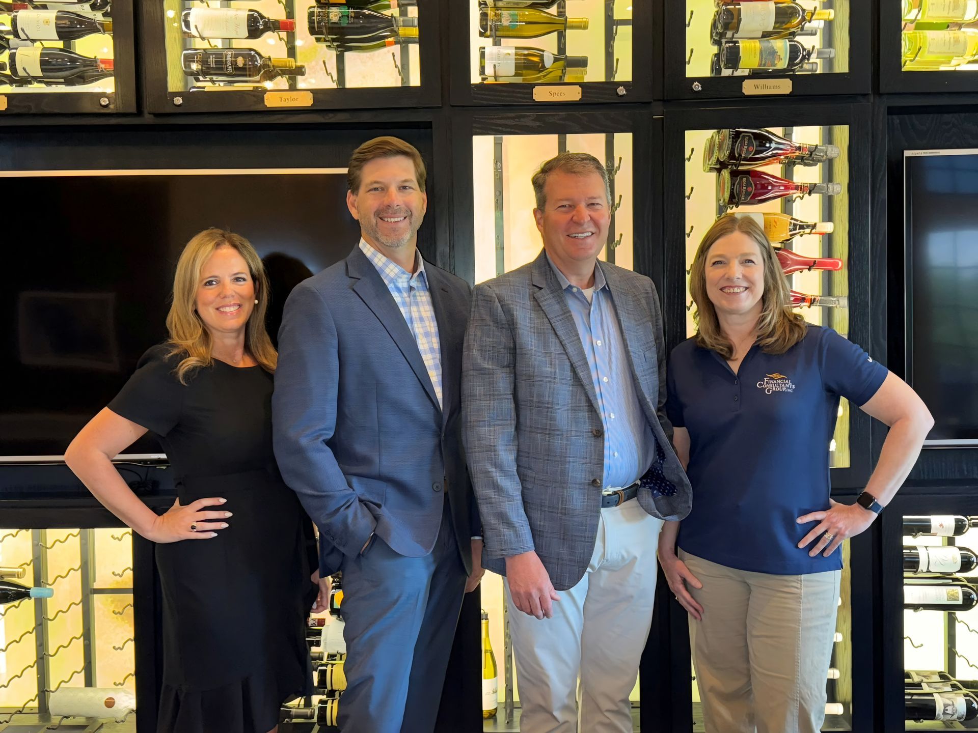 Four people pose for a photo in front of a wine rack. They are smiling, wearing professional attire.