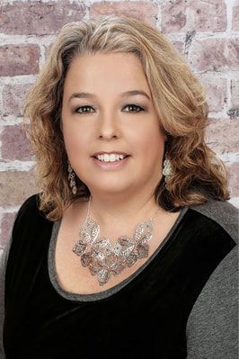 A woman wearing a necklace and earrings is smiling in front of a brick wall.