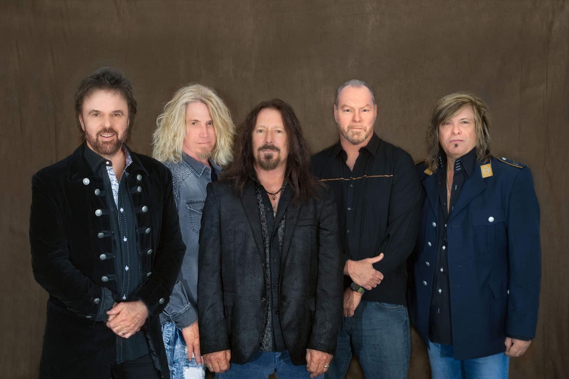 Five musicians posing together in dark jackets against a plain studio backdrop