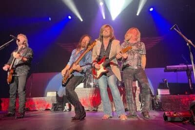 Rock band performing onstage under blue and white lights, with guitars and microphones.