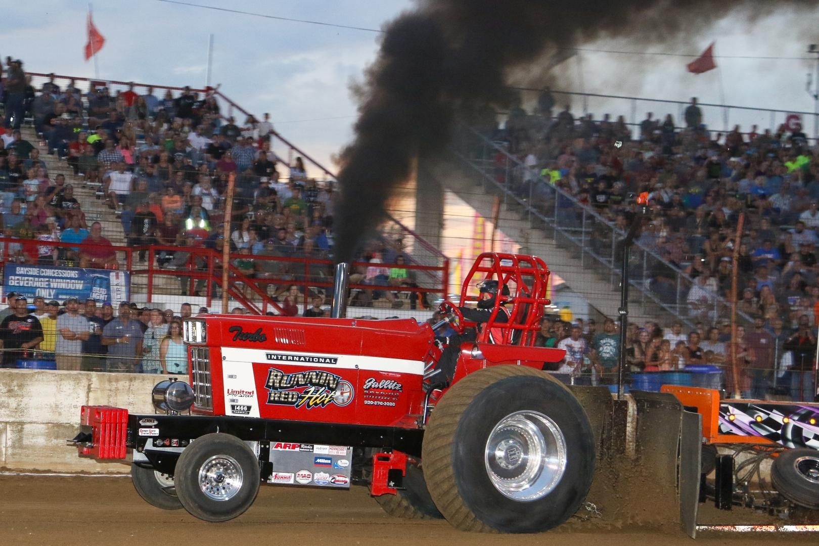 Red tractor-pull truck with black smoke in a dirt arena, crowd cheering in the stands.