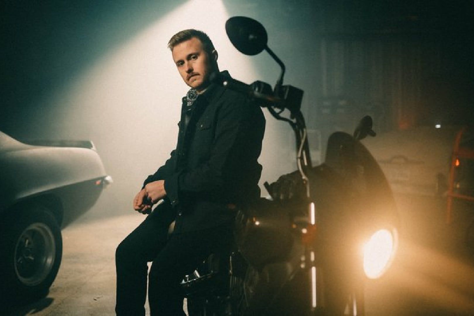 Man seated on a motorcycle at night, lit by a bright headlight in a smoky garage