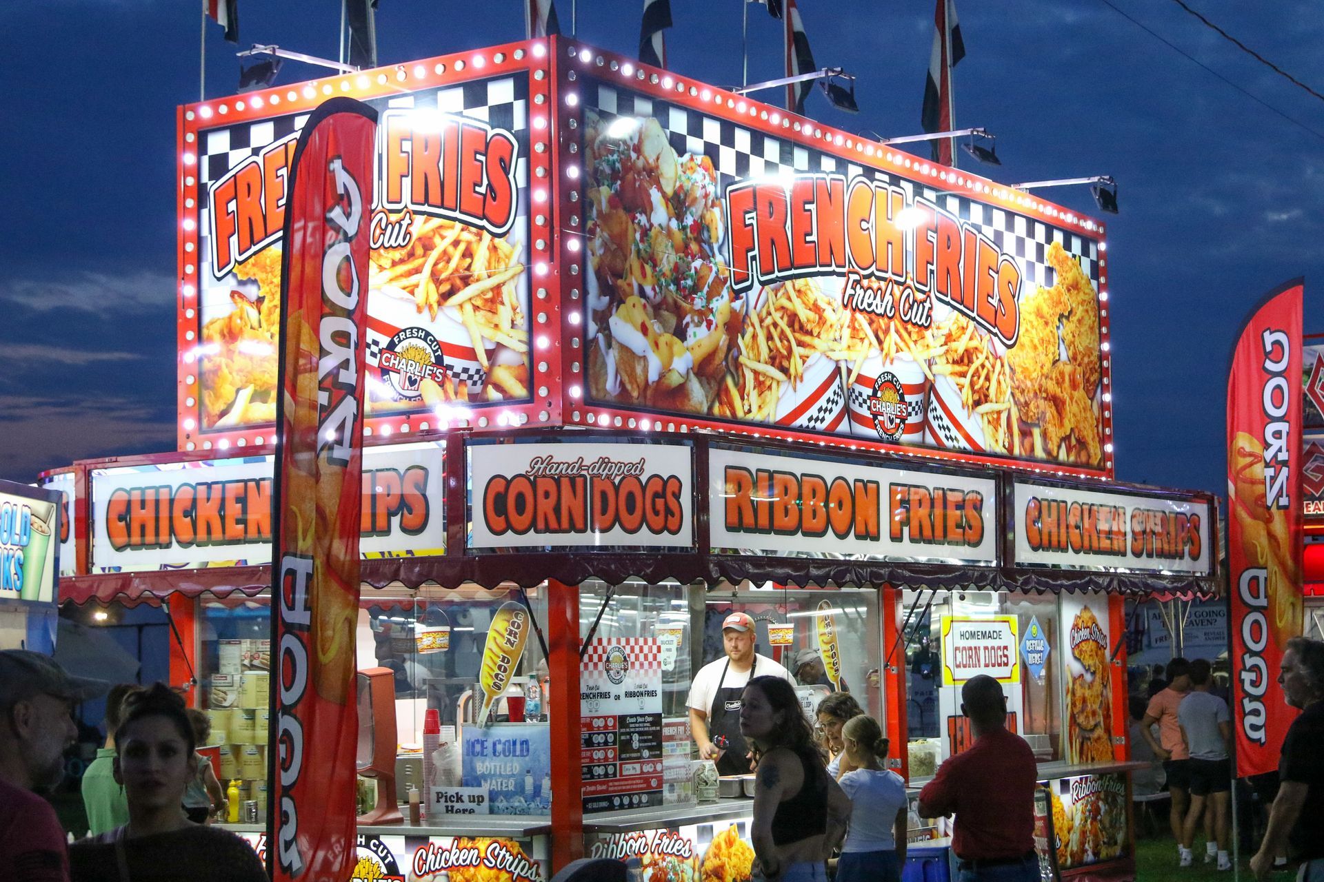 Brightly lit fair food stand with chicken fingers, corn dogs, and festival fries at dusk