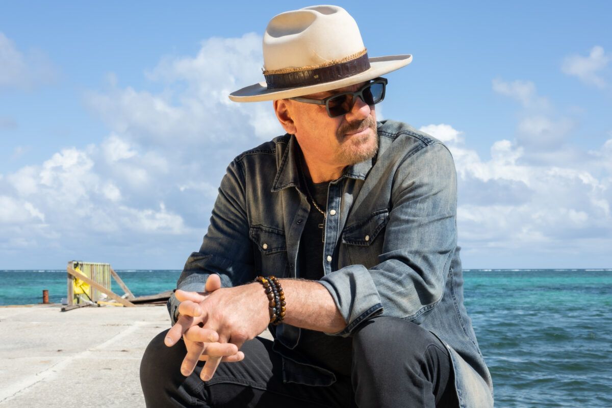 Man in a straw hat and sunglasses sitting on a beach with turquoise water and a bright sky behind him