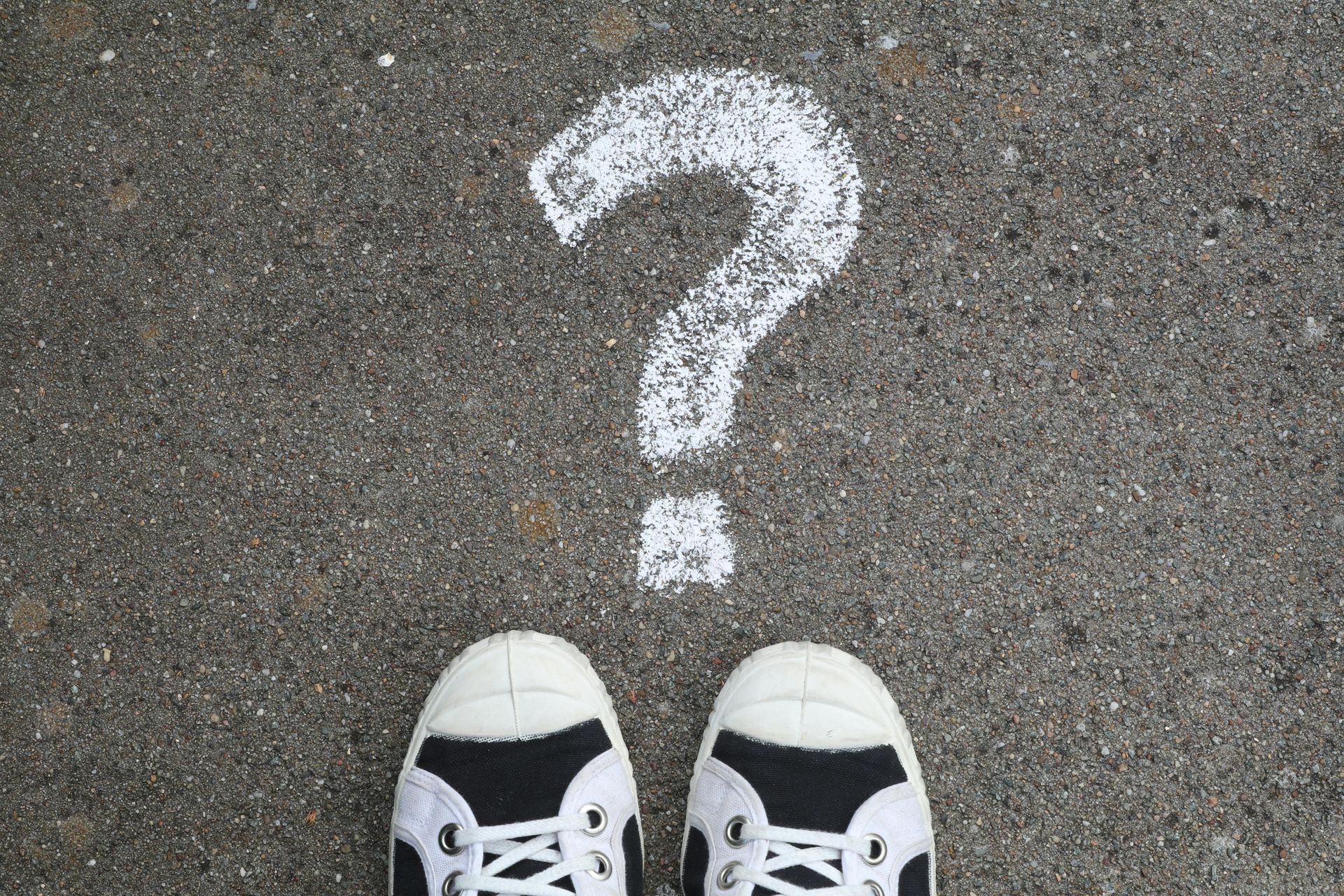 White sneakers standing beneath a white question mark spray-painted on asphalt.