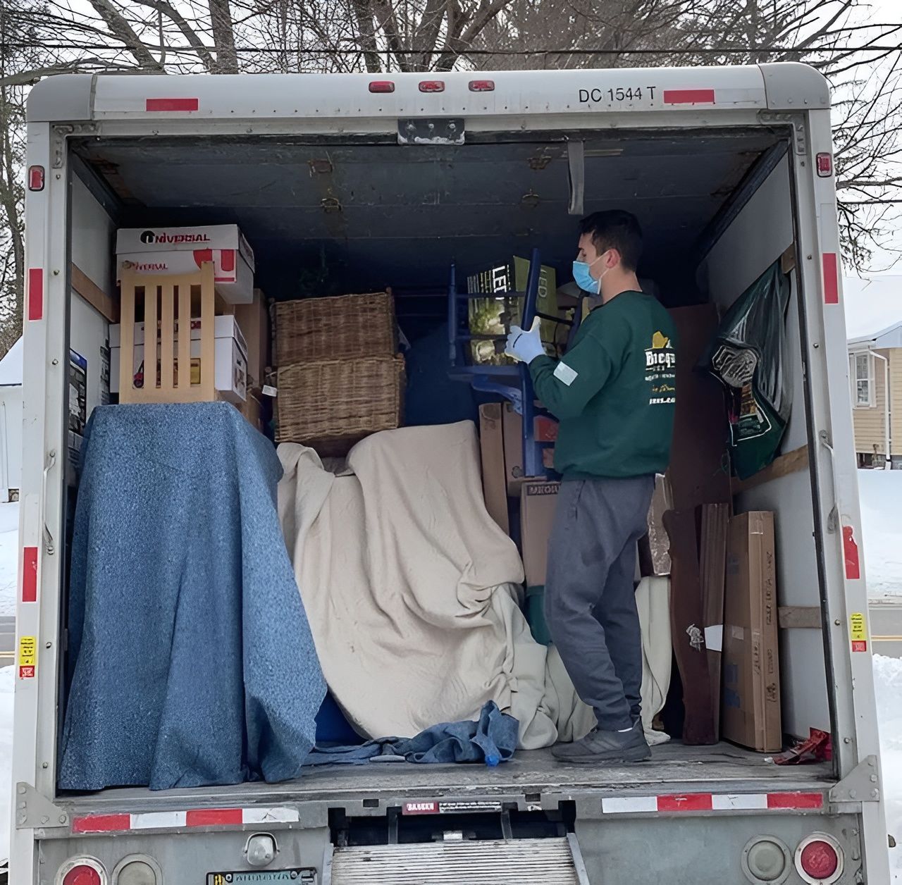 A person wearing a mask loads a moving truck with furniture, boxes, and a wicker basket. Snowy outdoors.