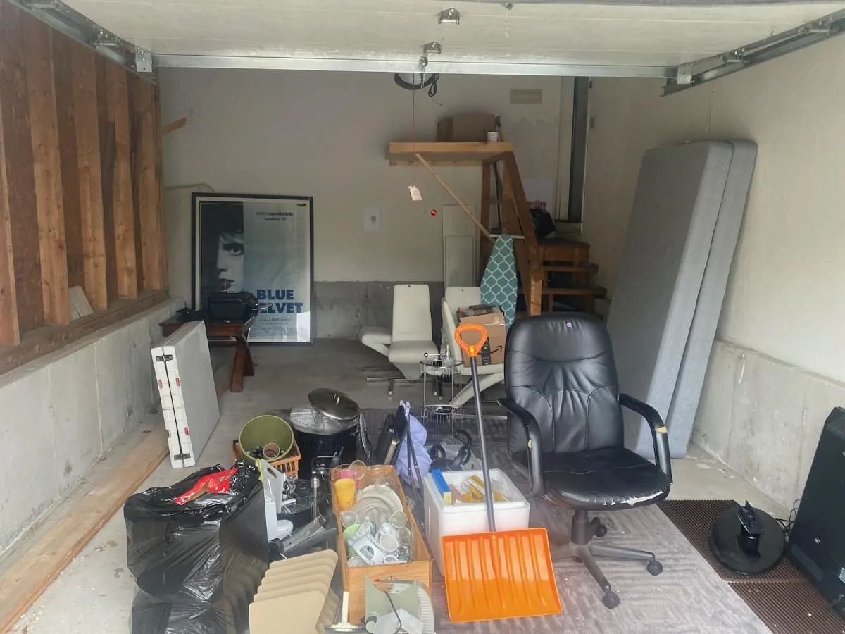 Cluttered garage with various items: tools, furniture, boxes, and trash. Beige walls, concrete floor, and open garage door.