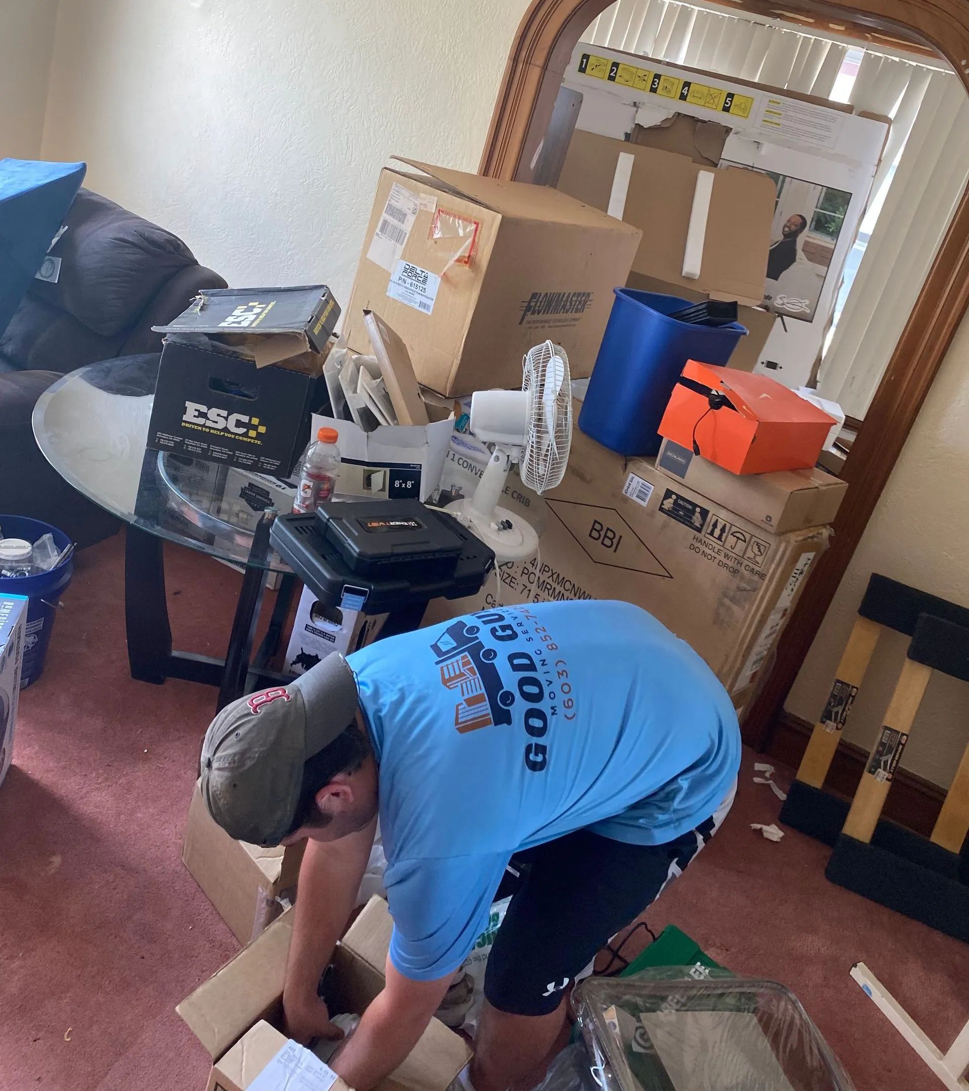 Person in blue shirt packing boxes in a cluttered room with furniture, mirror, and various items.