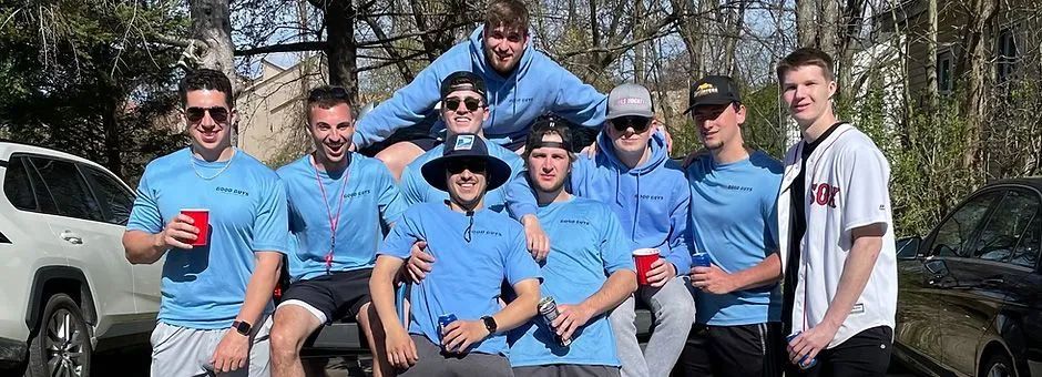 Group of young men posing outdoors; some wearing sunglasses, light blue shirts, holding cups; sunlight.