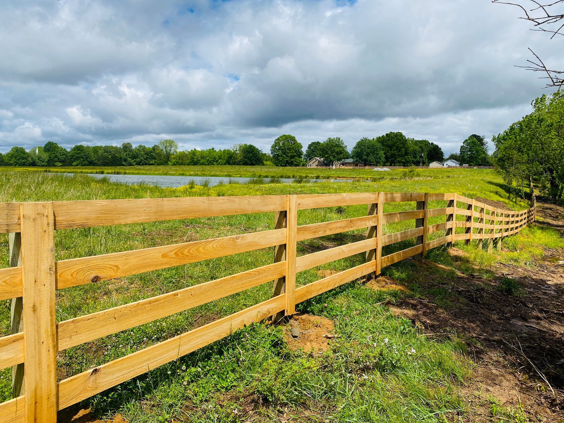 A wooden fence surrounds a grassy field with a pond in the background.