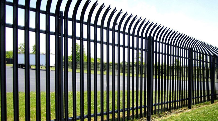 A black metal security fence with curved, spiked tops, bordering a grassy area and an asphalt lot with a truck trailer.