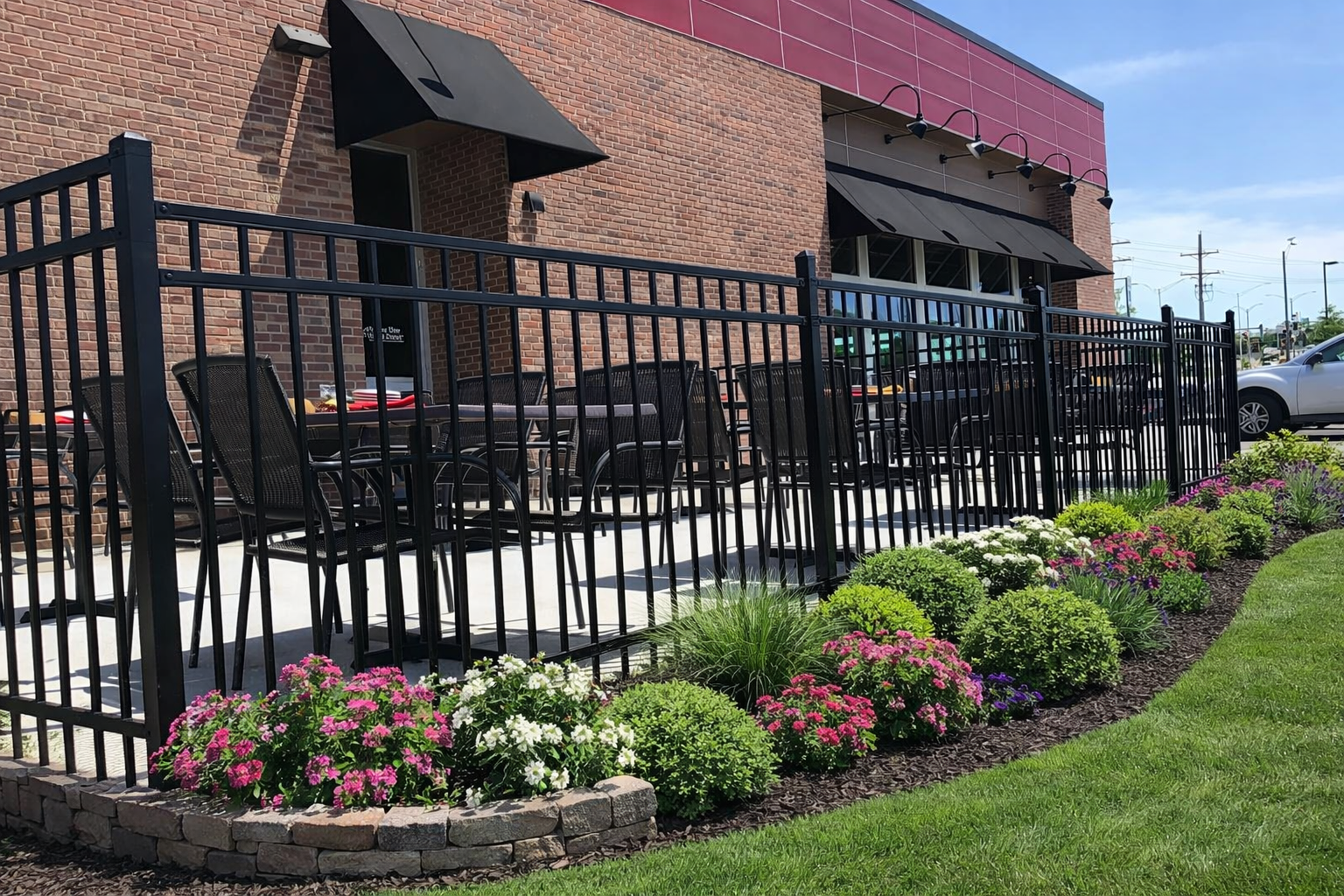 A brick building features an outdoor dining patio enclosed by a black metal fence, bordered by shrubs and flowers.