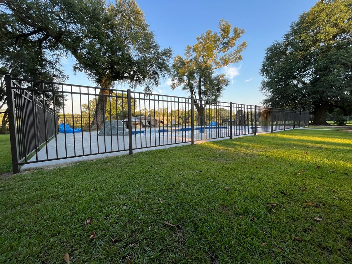 Black fence encloses a pool area with a blue pool on a grassy lawn, trees in the background.