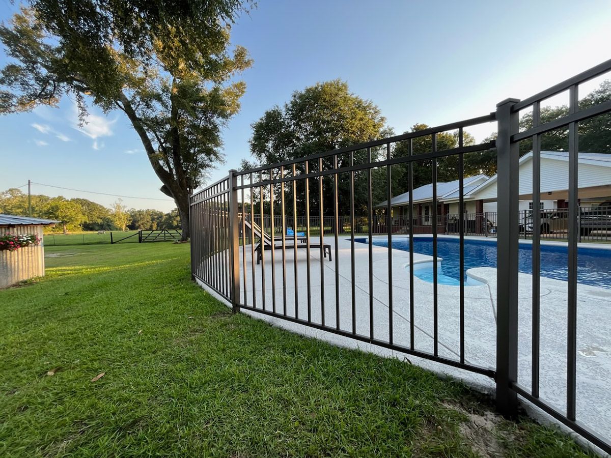 Black fence surrounds a backyard pool, with trees and a house in the background under a blue sky.