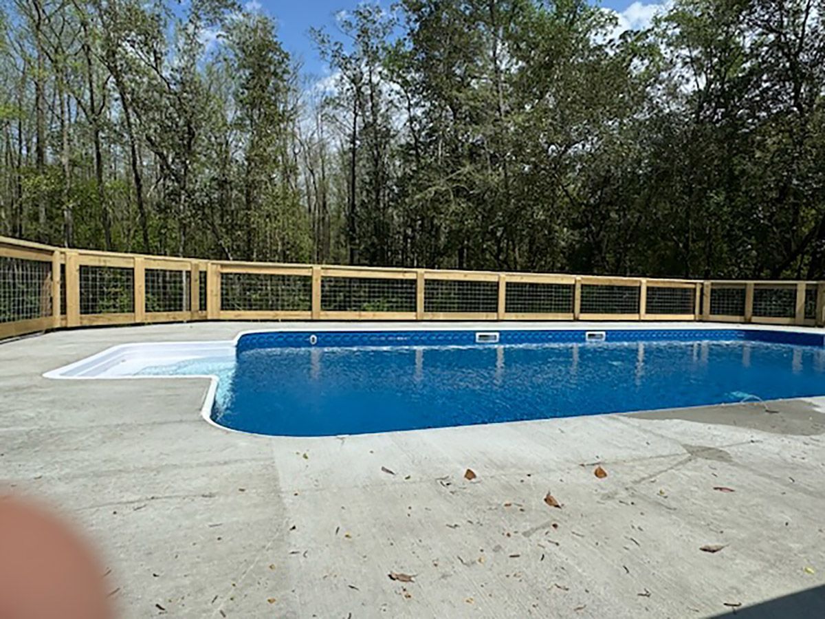 Swimming pool with concrete deck, surrounded by a wooden fence and trees in the background.