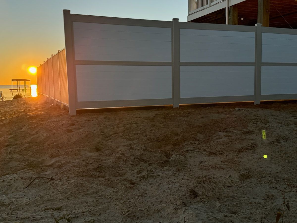 Sunset over water, viewed through a white fence on a sandy beach.