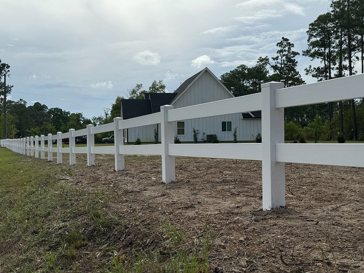 A white fence surrounds a dirt field with a house in the background.