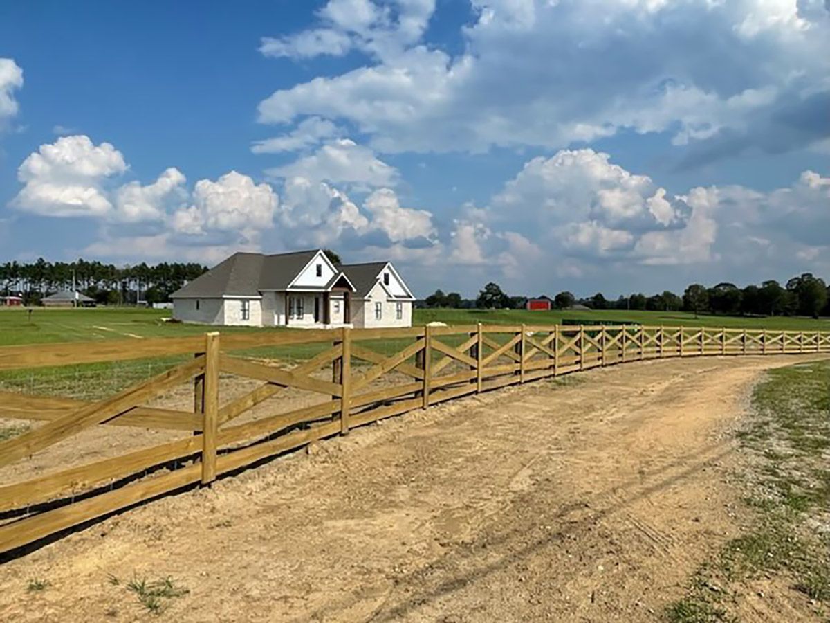 A wooden fence surrounds a dirt road and a house in the background.