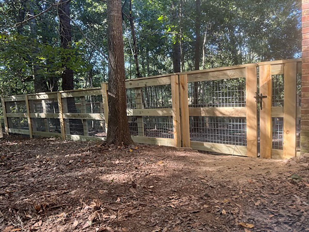 Wooden fence with wire mesh, built in a wooded area. A gate is open, framed by a brick pillar.