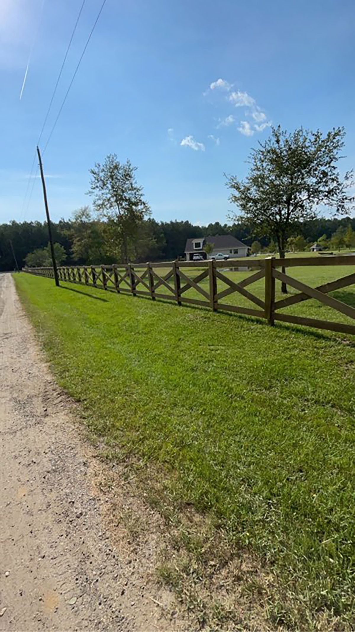 A wooden fence borders a grassy field with a house in the distance under a clear blue sky.