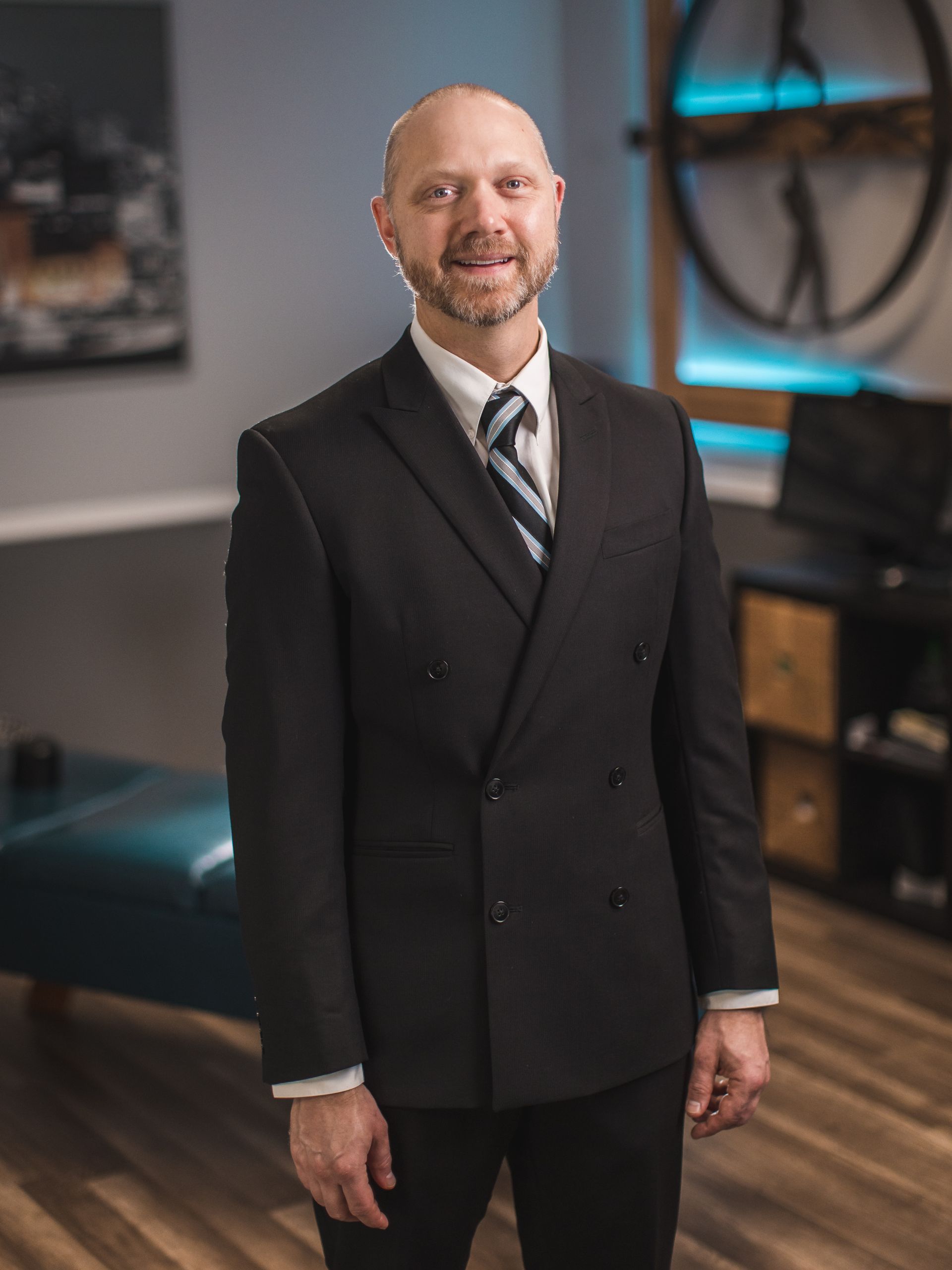 Man in black suit and striped tie smiles, standing in an office.