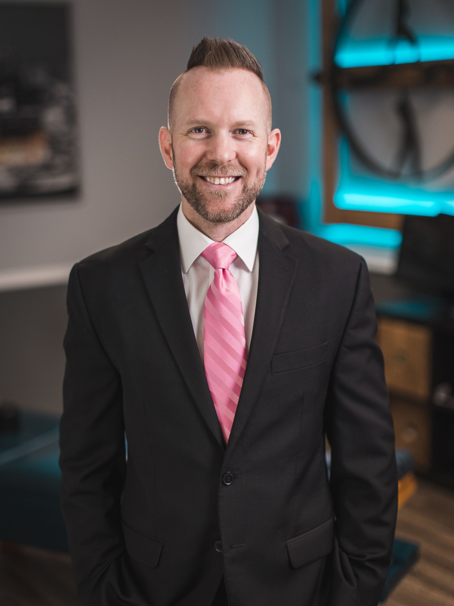 Man in black suit and pink tie smiles, standing in an office.