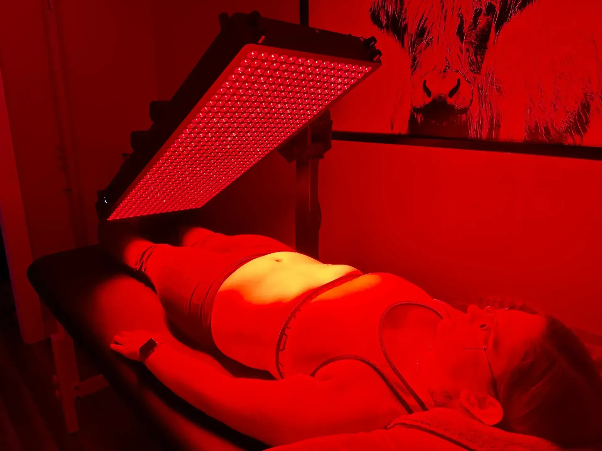 Person lying under a red light therapy panel; setting appears to be a treatment room.