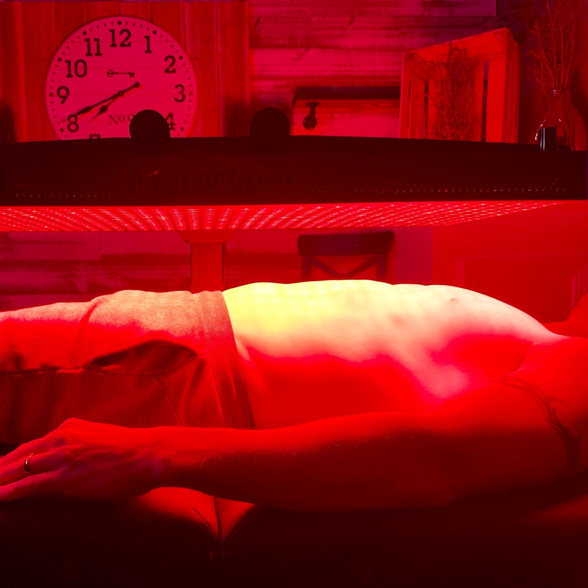 Person receiving red light therapy on their back, lying down. A clock and wooden elements are in the background.