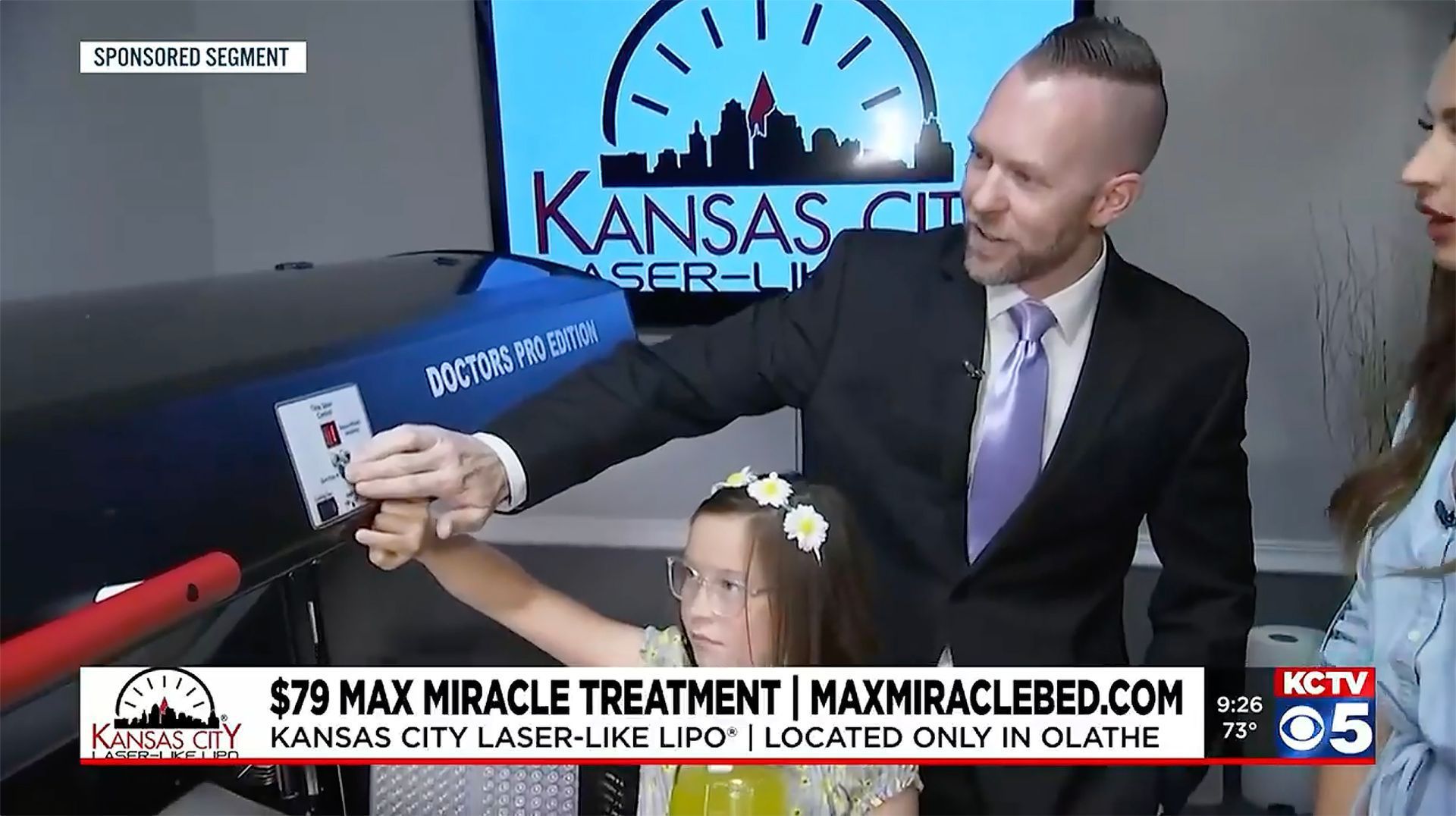 Man with a purple tie holding a device with a child and woman on a TV set in Kansas City.