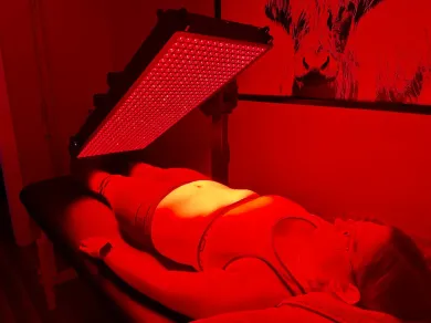 Person receiving red light therapy on stomach, lying on a massage table. Red light shines from overhead panel.