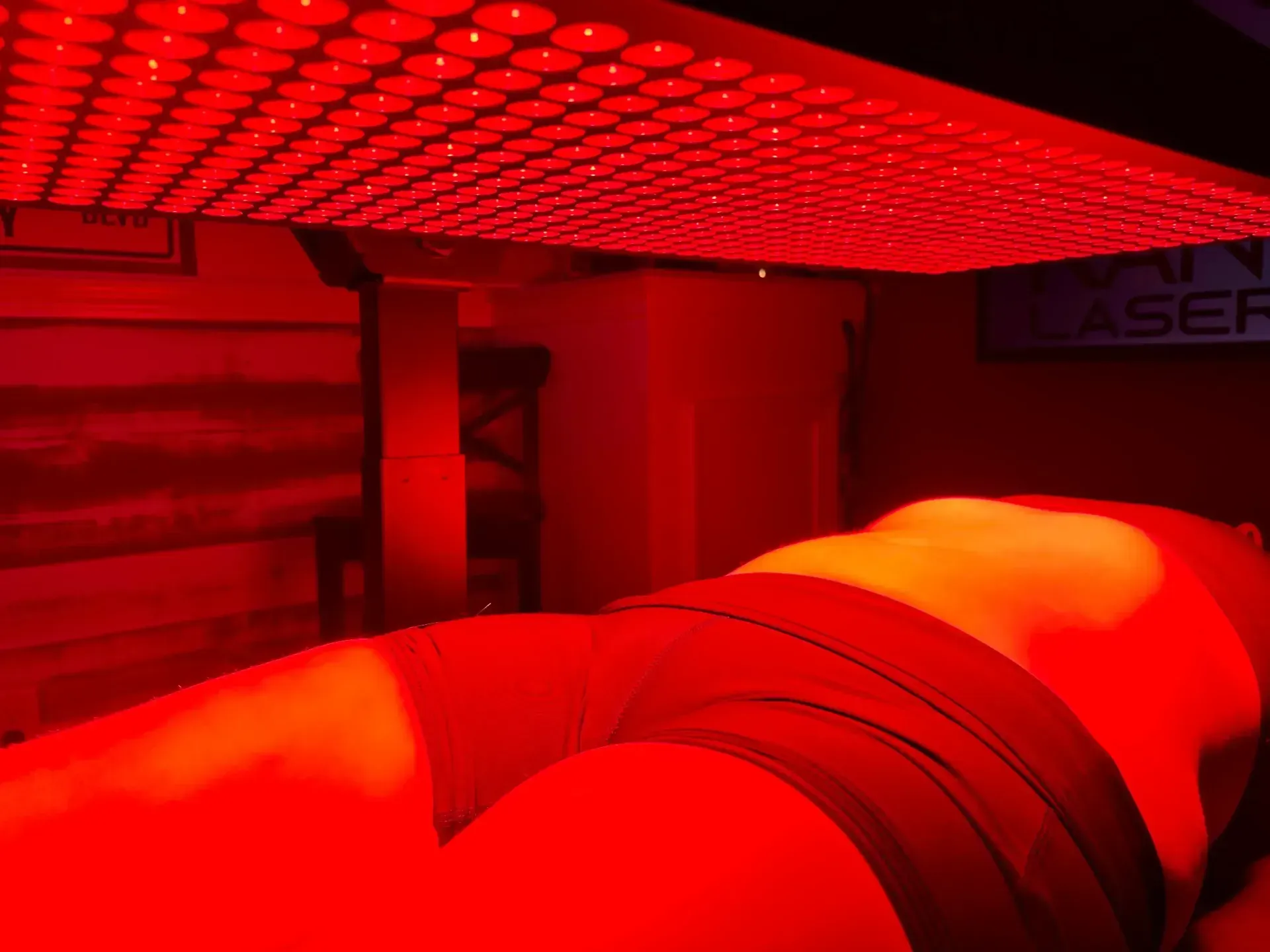 A person lies under a red light therapy panel in a dimly lit room.