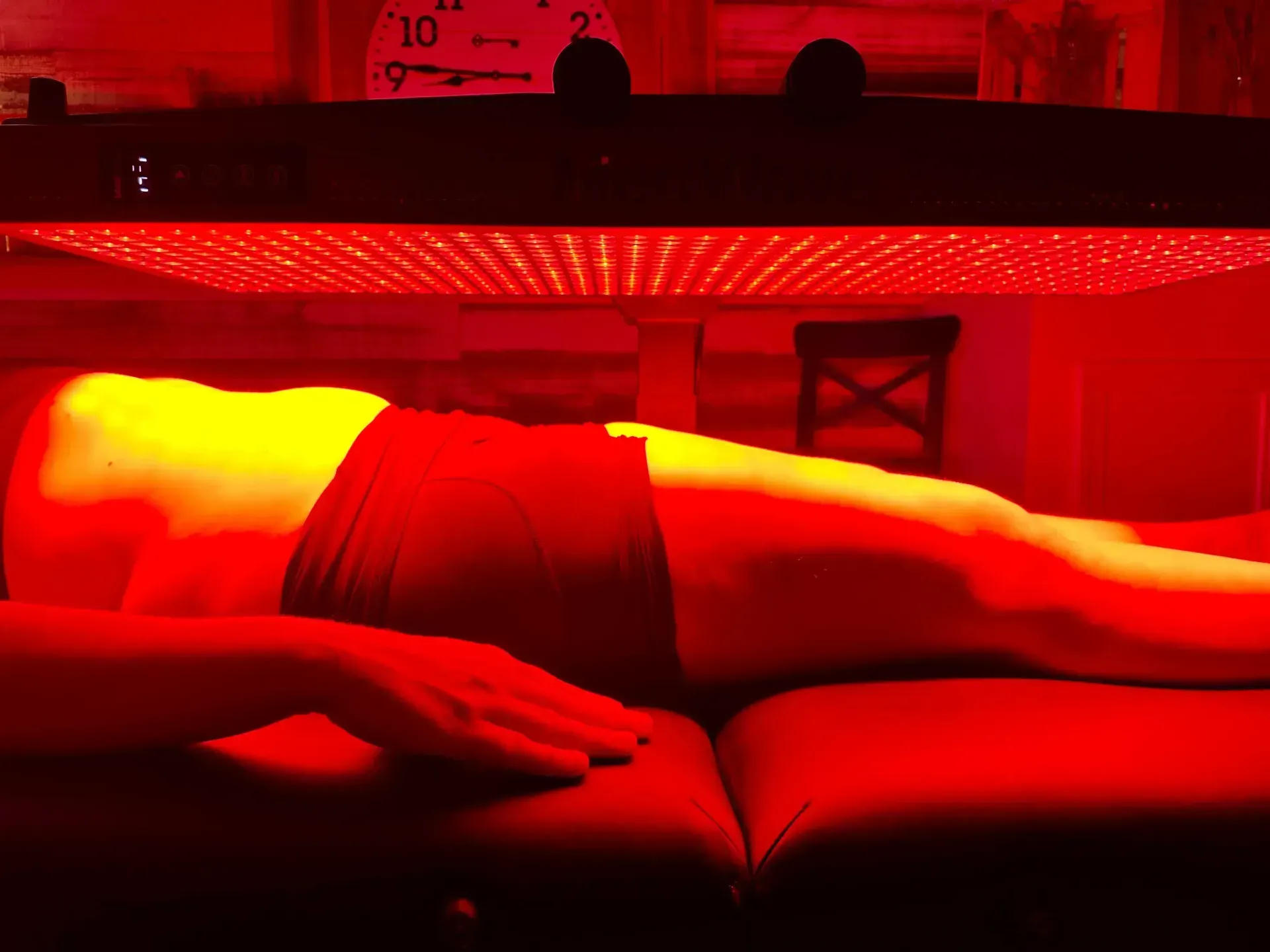 Person lies under a red light therapy panel. Red light bathes the body, illuminating the panel above.
