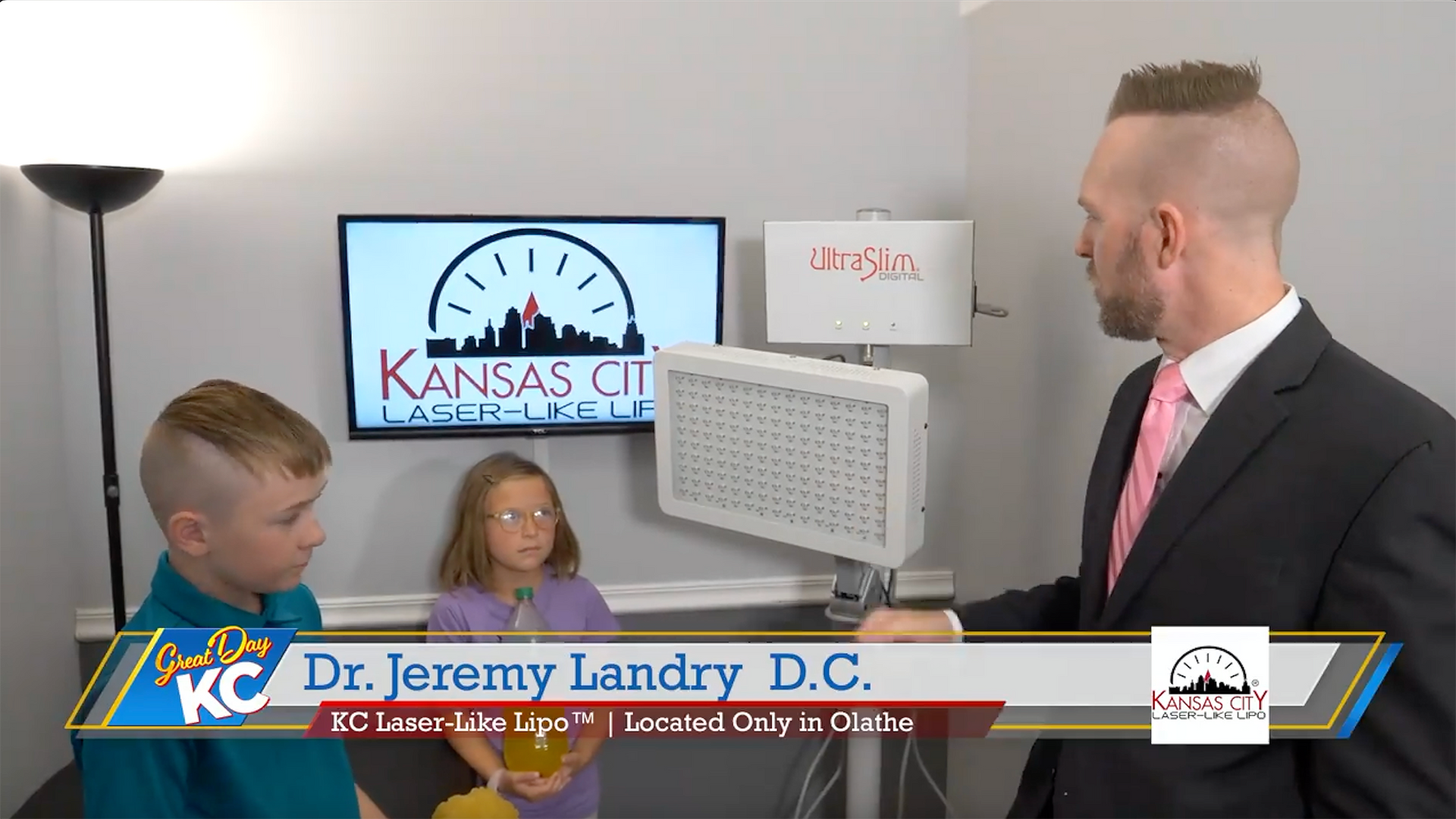 A man in a suit points at a light device while two children watch. The business logo is in the background.