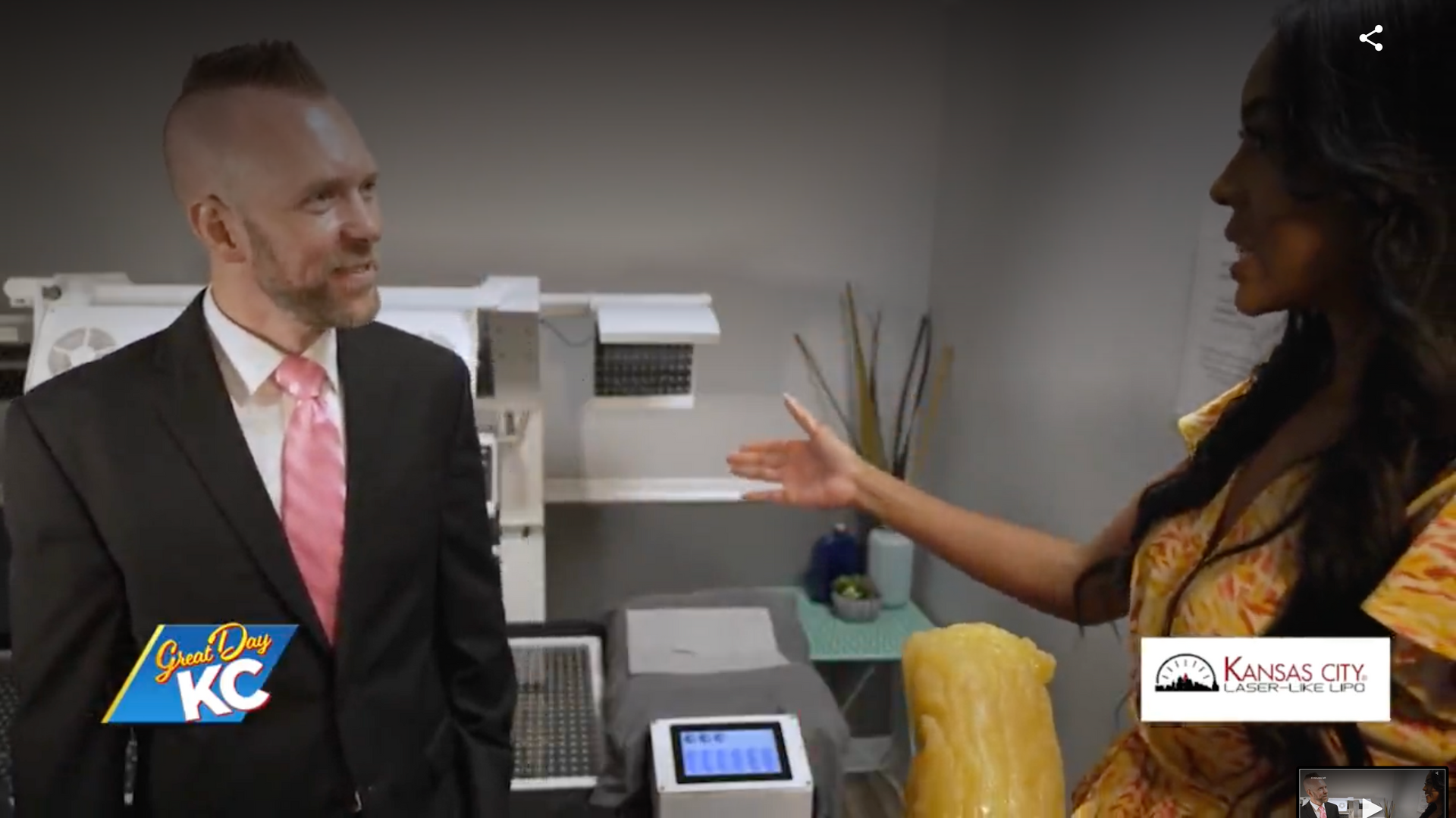 Man in suit and woman in yellow dress talking in a medical office, gesturing towards equipment.