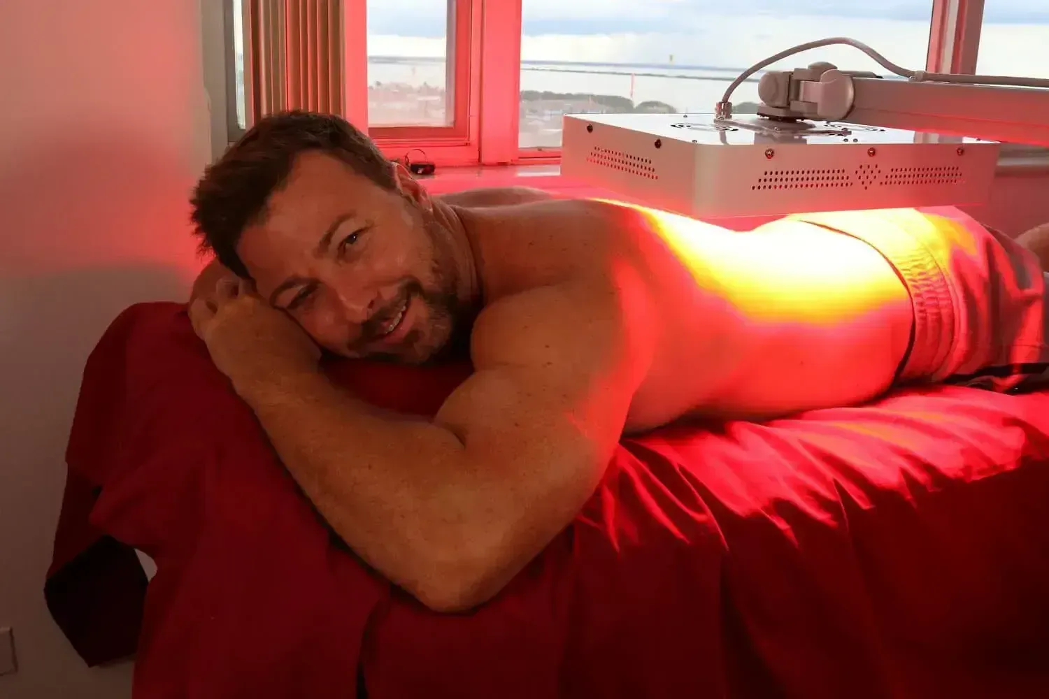 Hombre recibiendo tratamiento de terapia de luz roja, sonriendo, acostado en una mesa de masajes cerca de una ventana.