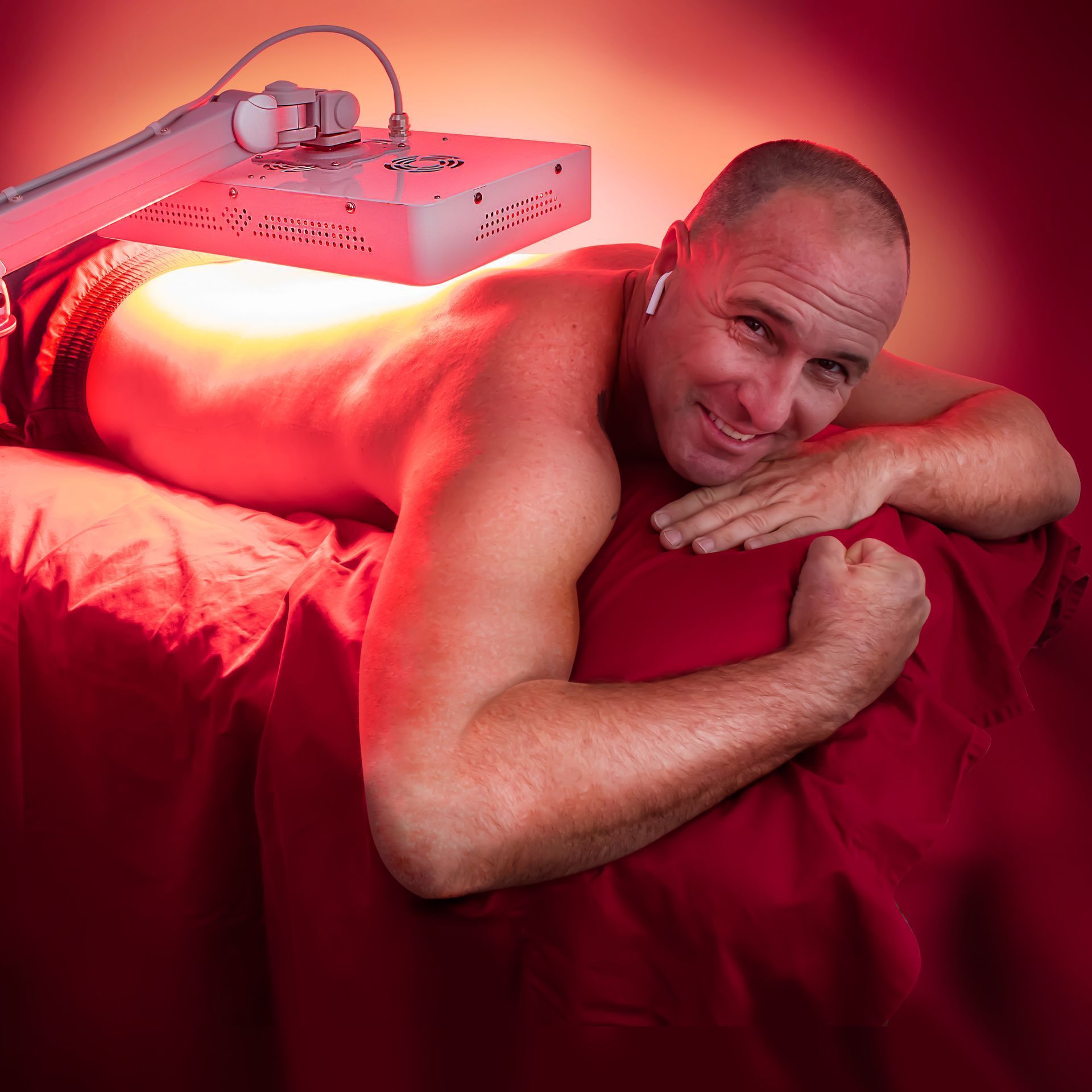 Man lies on red surface under red light therapy lamp, smiling and flexing bicep.