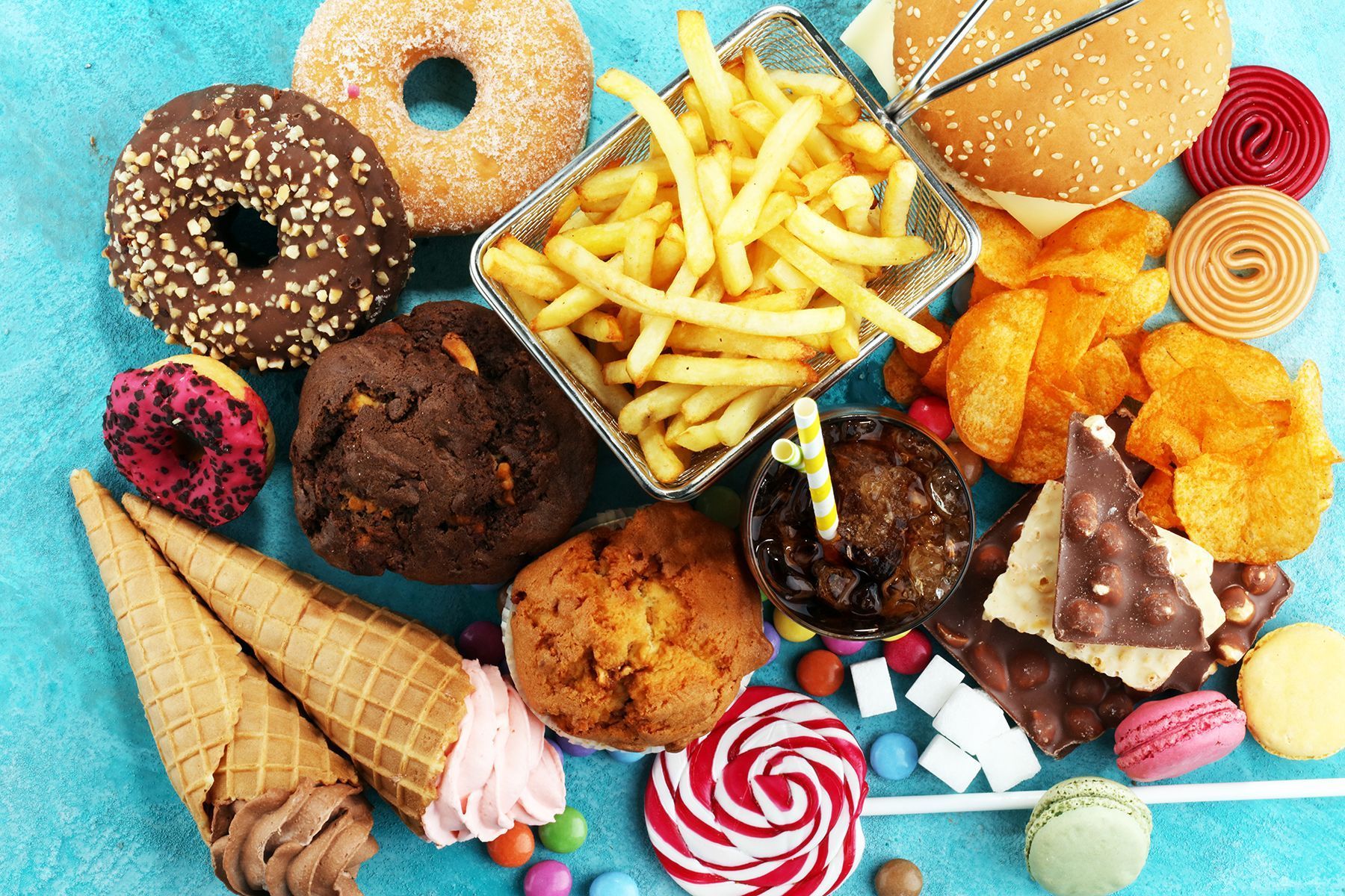 Assorted unhealthy foods: donuts, fries, burger, chips, ice cream, candy, muffins, cookies, on a blue surface.