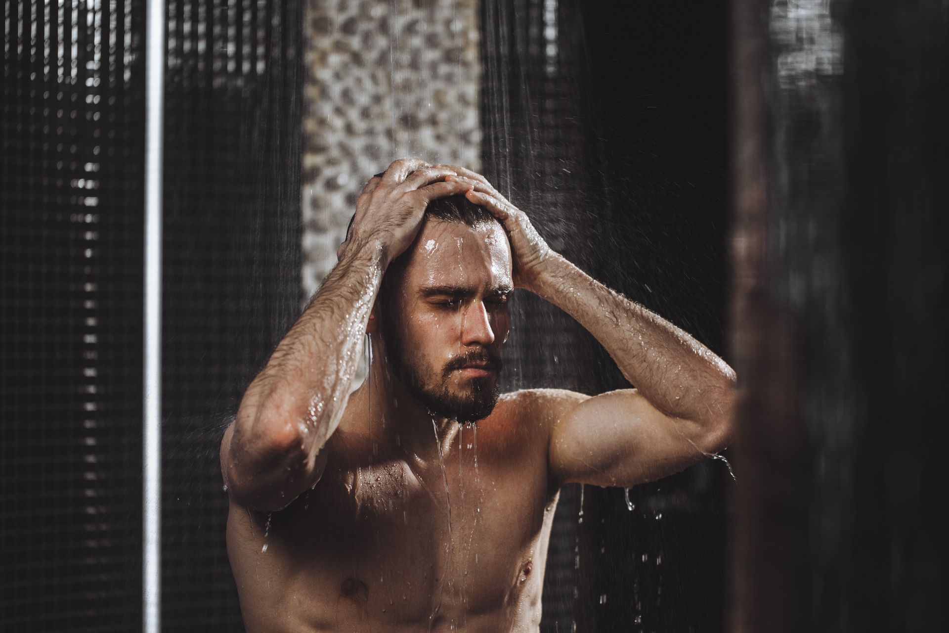 Man in shower, hands in hair, water streaming. Dark tiled backdrop.