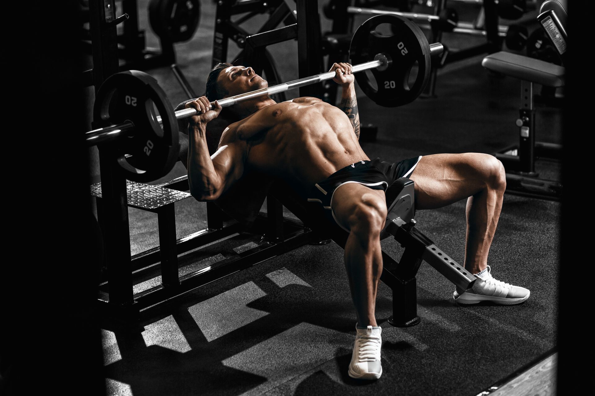 Man bench pressing barbell in a gym, with muscular physique, lying on a bench, legs extended.