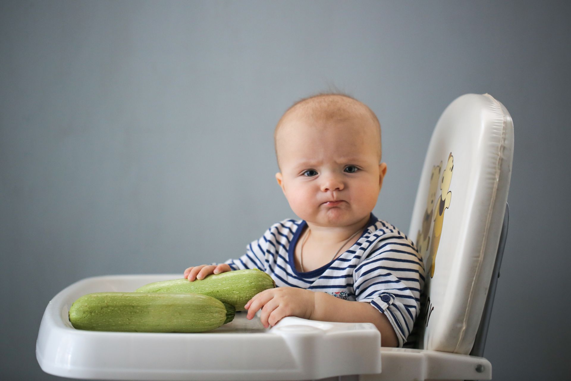 Baby in high chair with frown holding two zucchinis.