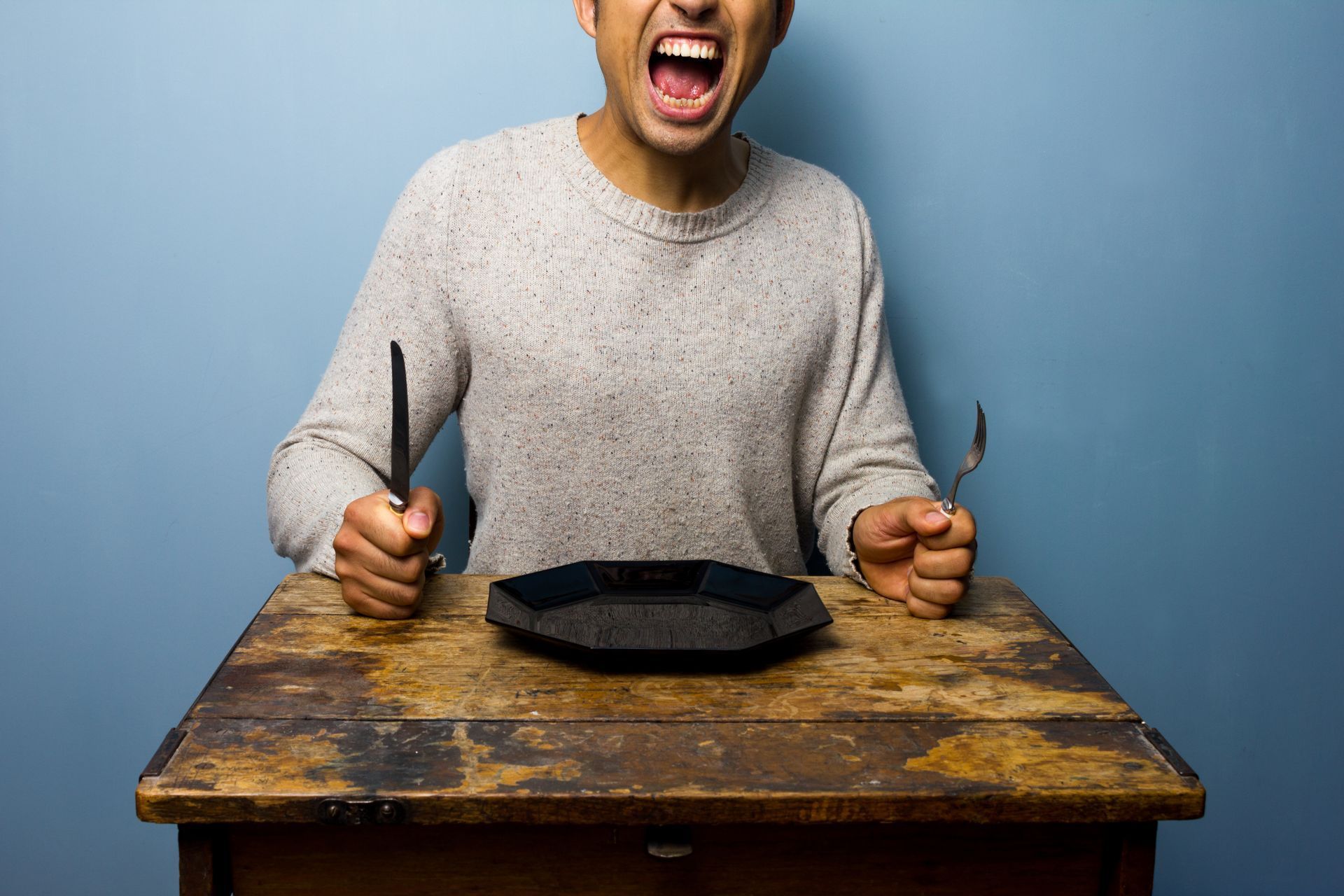 Person yelling at an empty plate, holding silverware, sitting at a wooden table against a blue wall.