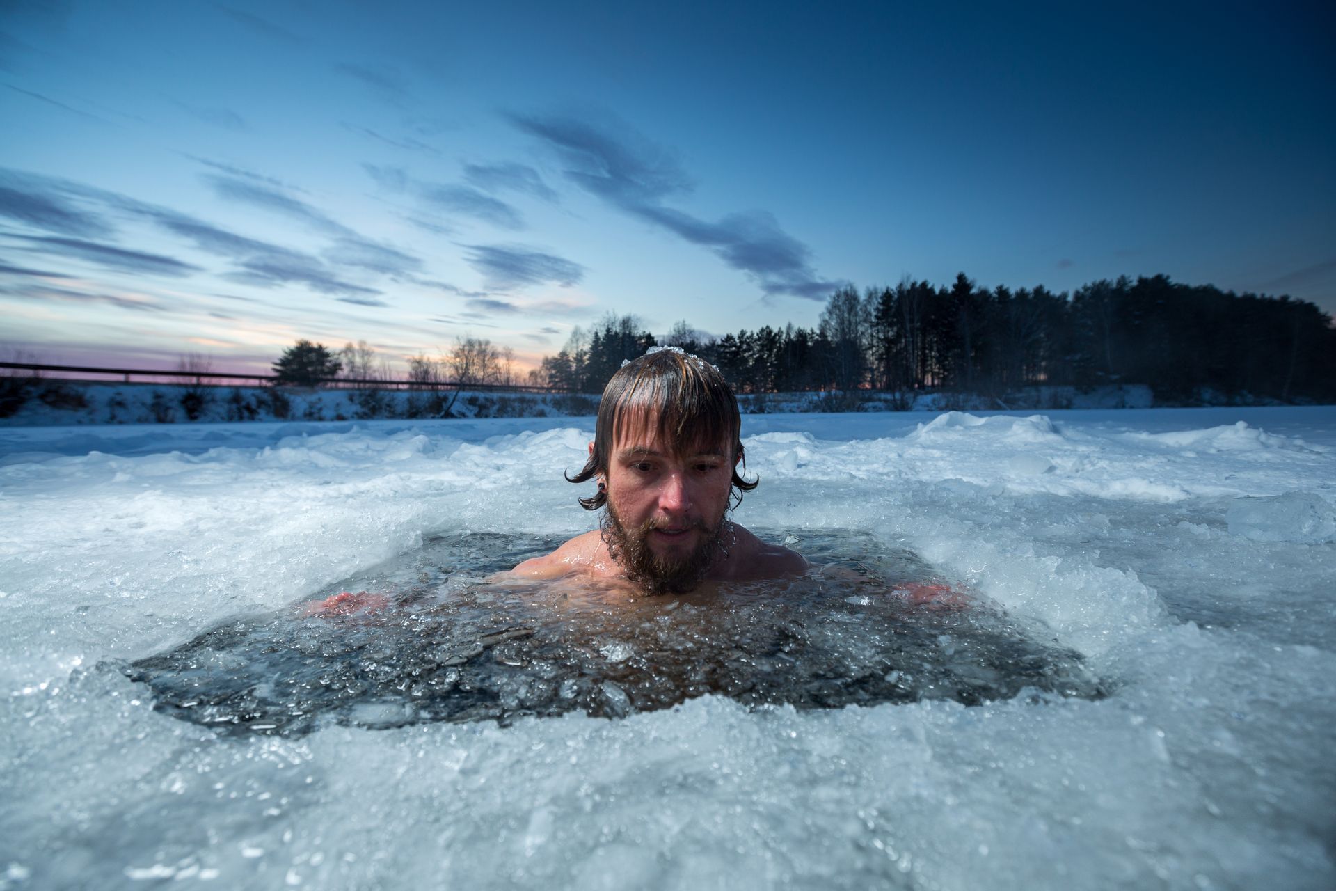 Man submerged in icy water, looking ahead, ice surrounding him. Dusk setting over trees and a snow-covered landscape.