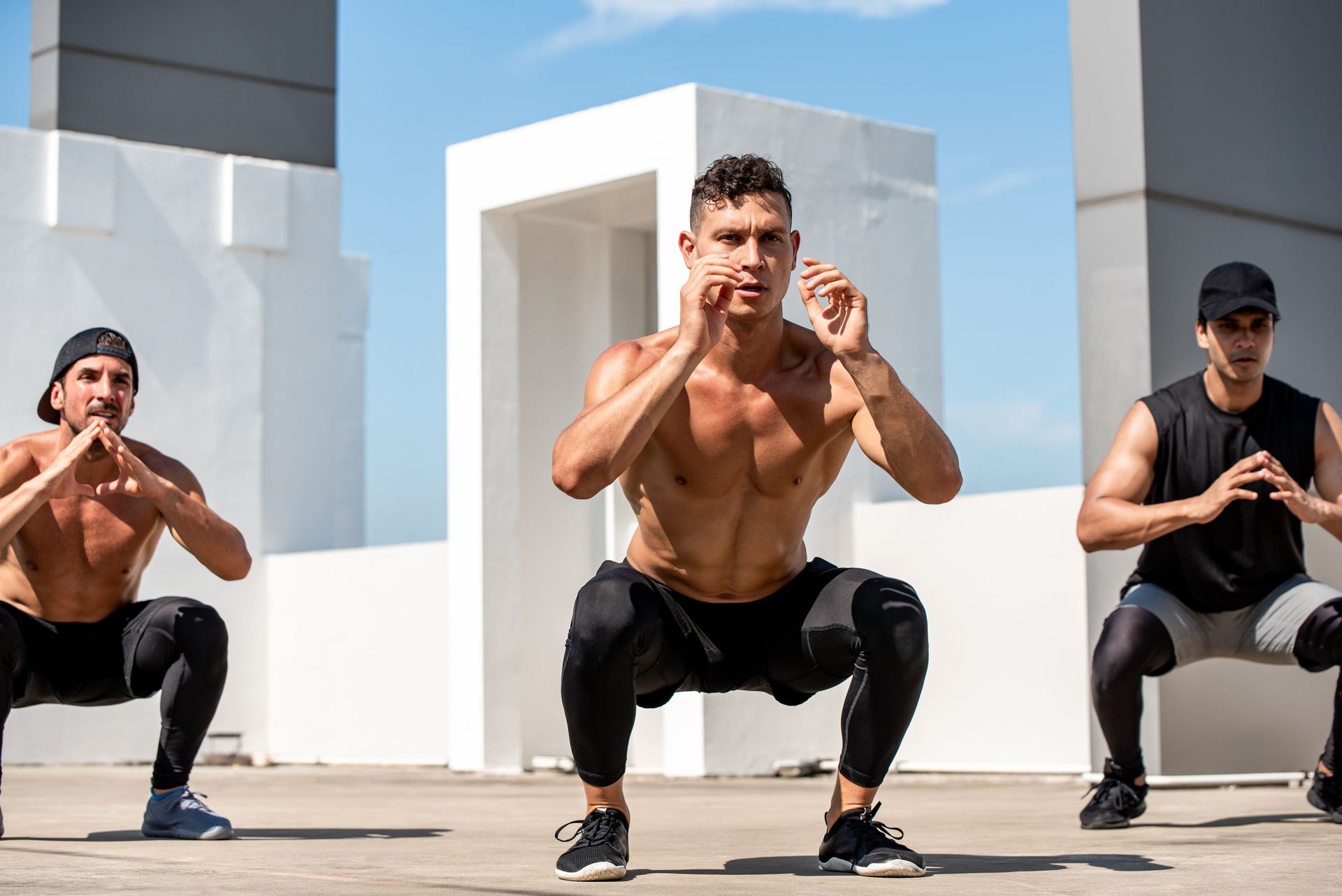 Three men doing squats outdoors on a rooftop; one is shirtless, all are wearing athletic clothing.